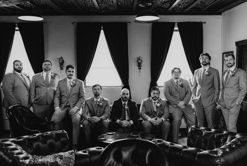 Black and white photo of groomsmen in suits posing on leather couches in an elegant room before a wedding.