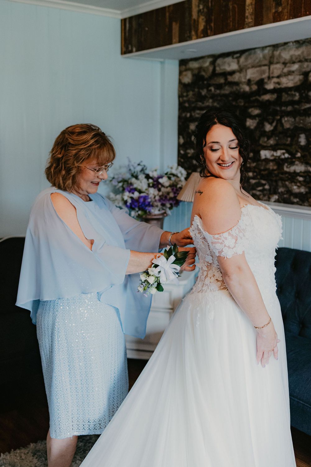 A bride in a white lace gown smiles as an older woman in blue helps her with a floral accessory before the wedding.