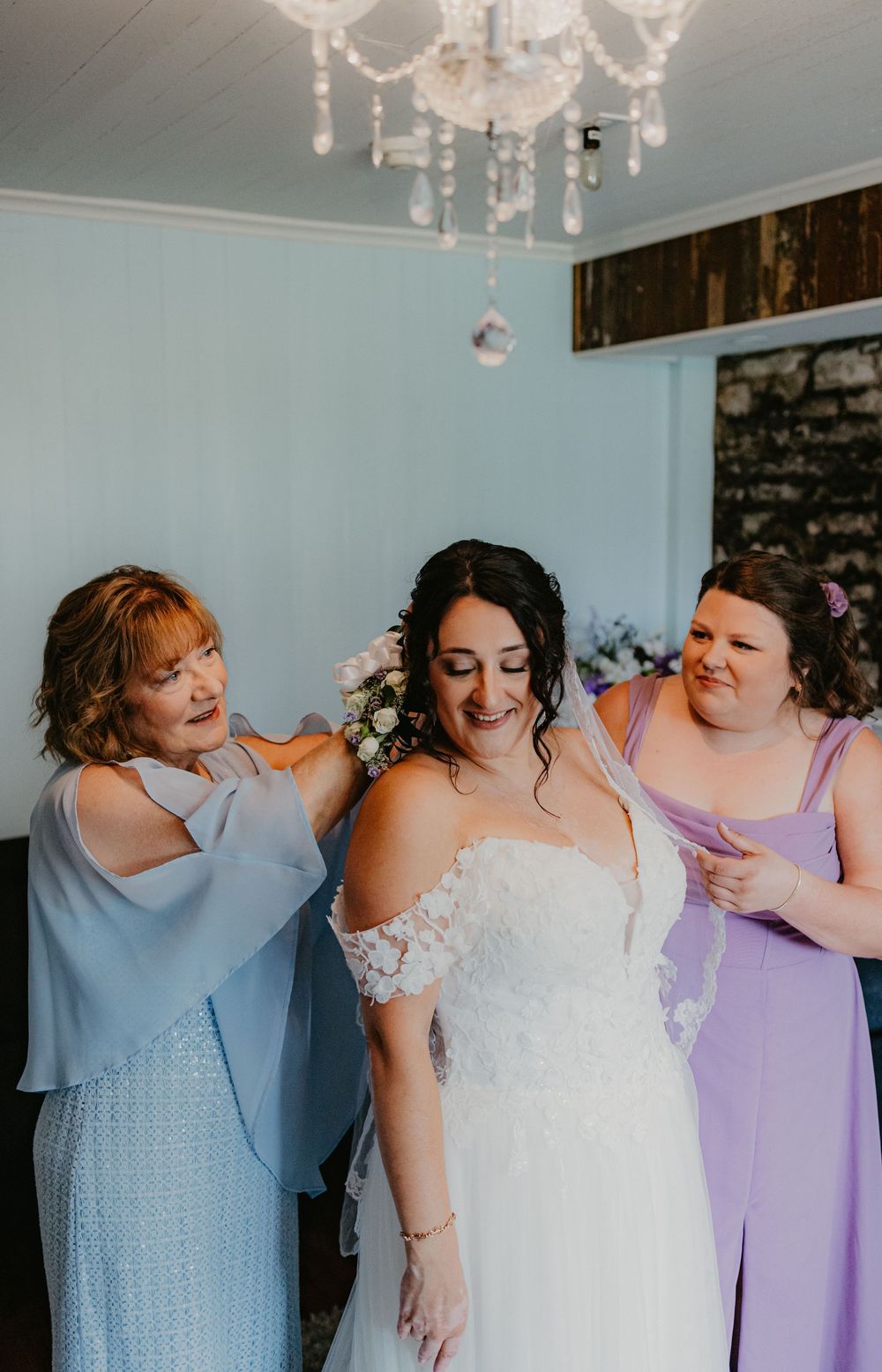 Bride in white lace gown being helped by mother in blue and bridesmaid in lavender dress under crystal chandelier.