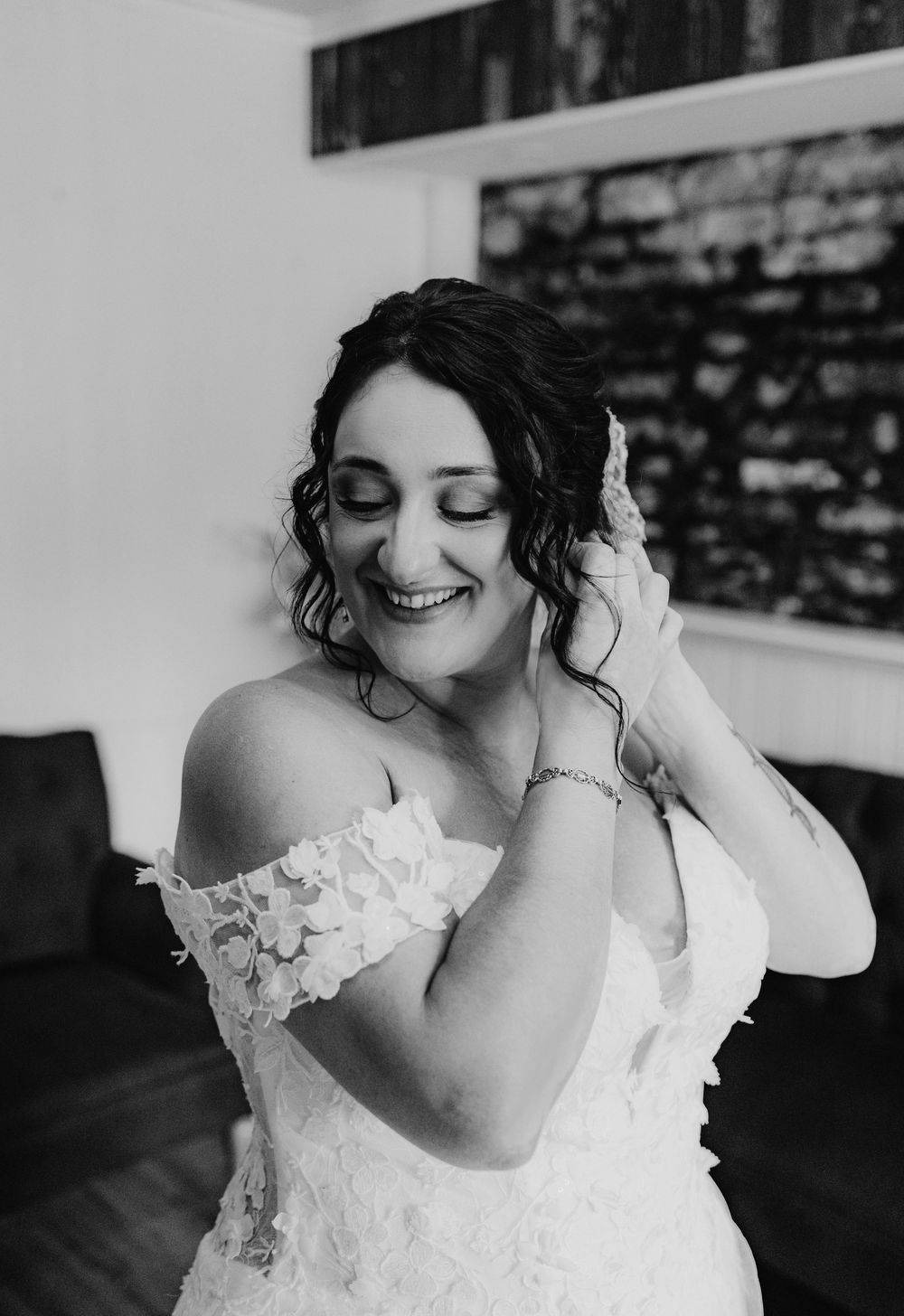 Black and white photo of a smiling bride in an off-shoulder lace gown adjusting her earring while getting ready.