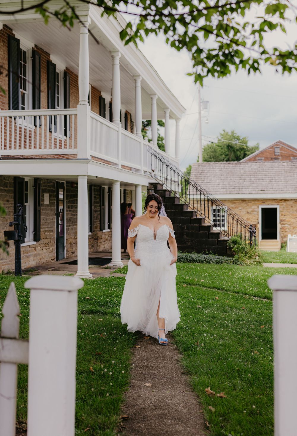 Bride in off-shoulder white wedding gown walking along garden path beside historic brick building with white columns.