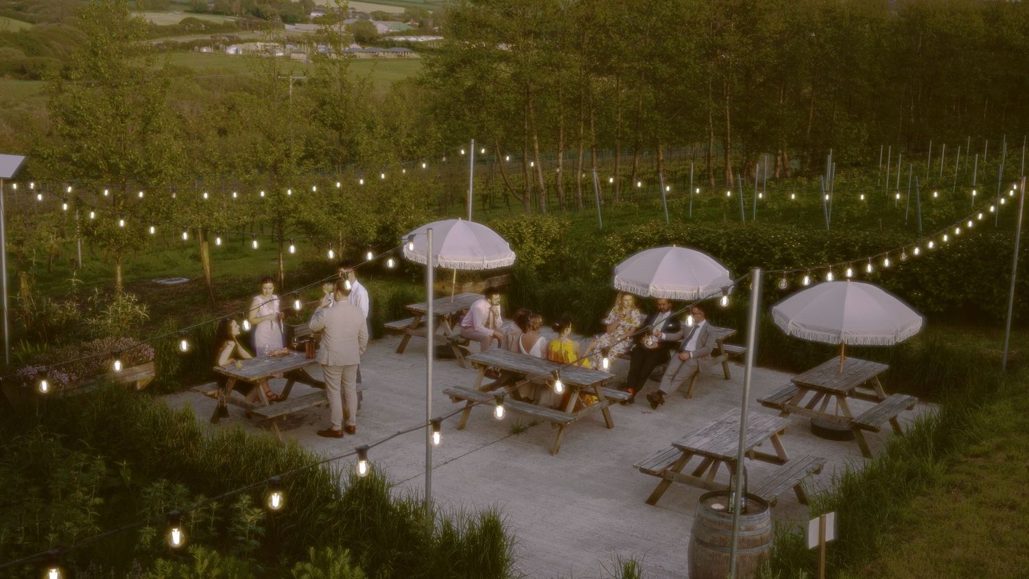 Outdoor patio with string lights, umbrella tables, and guests dining at dusk surrounded by lush greenery.