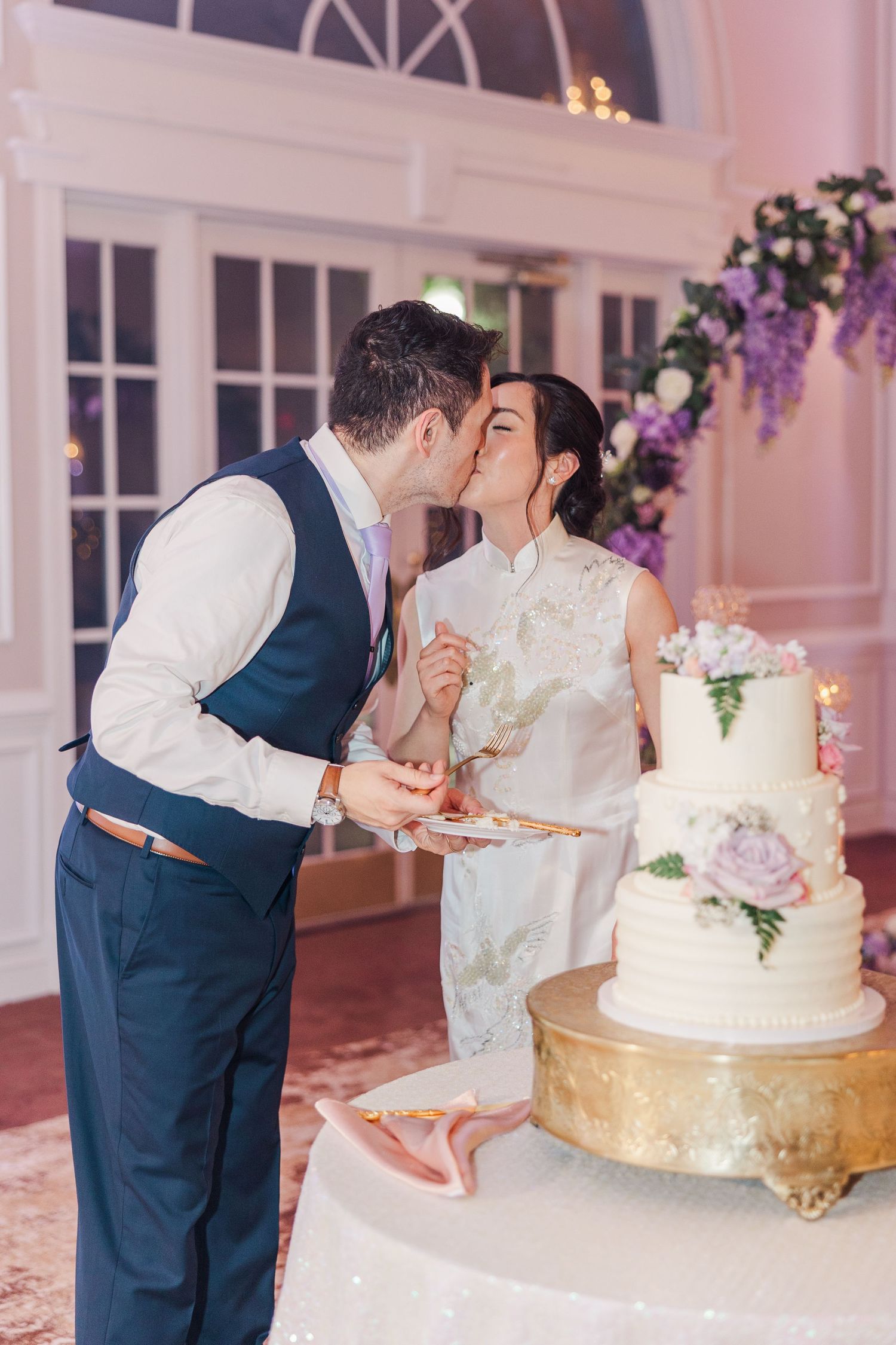 Bride and groom share a kiss while cutting their elegant white tiered wedding cake at their reception.