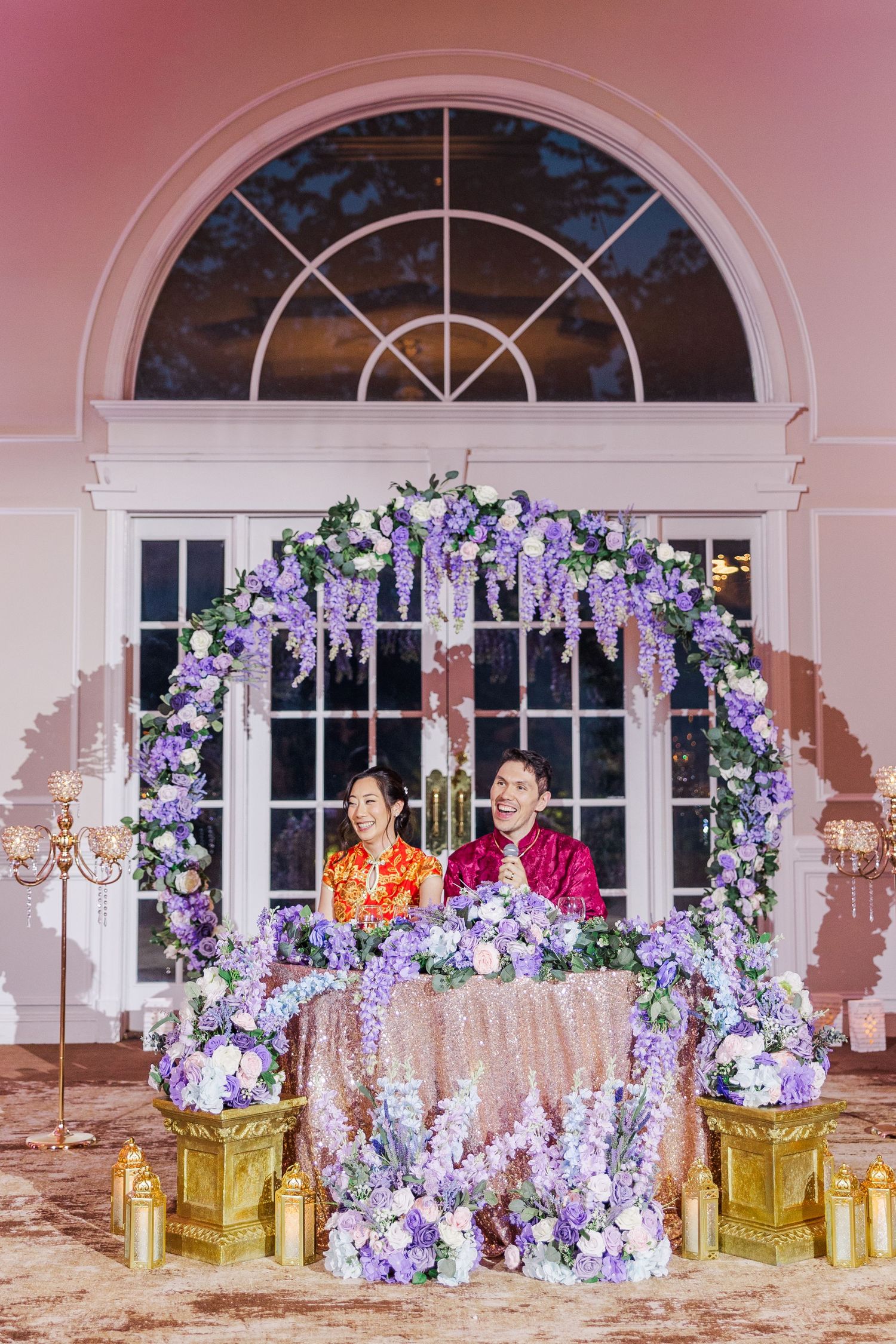 Couple in traditional Chinese wedding attire seated at floral arch with purple wisteria blooms and candles at elegant venue.