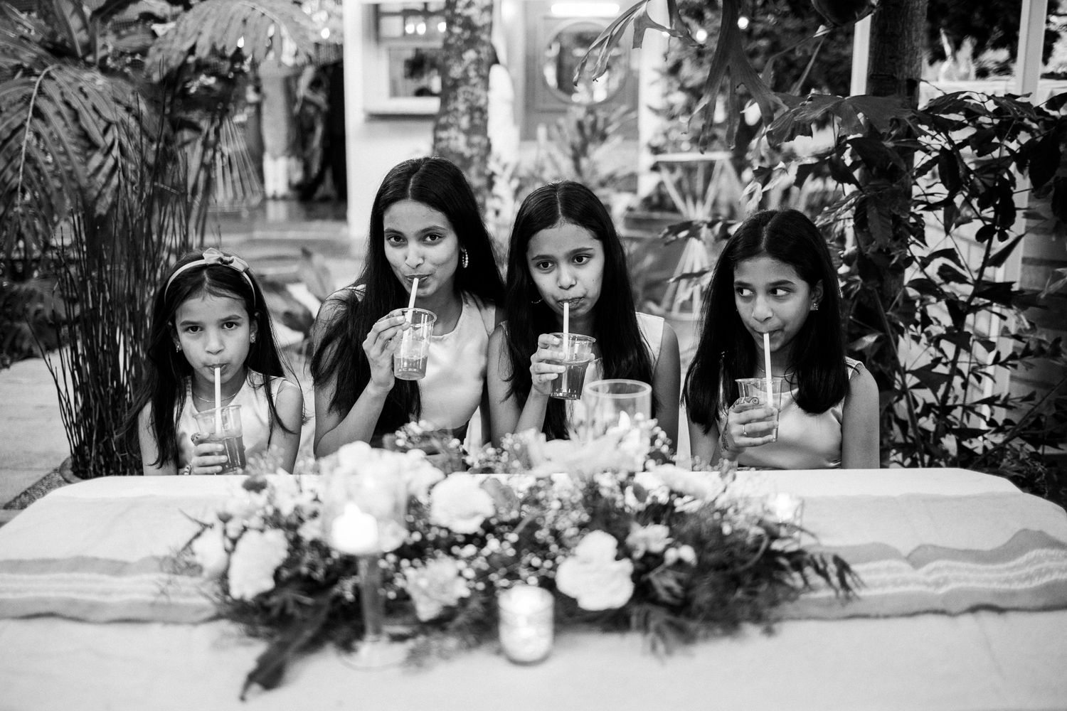 Four young girls in white dresses sip drinks through straws at a floral-decorated wedding table in black and white photo.