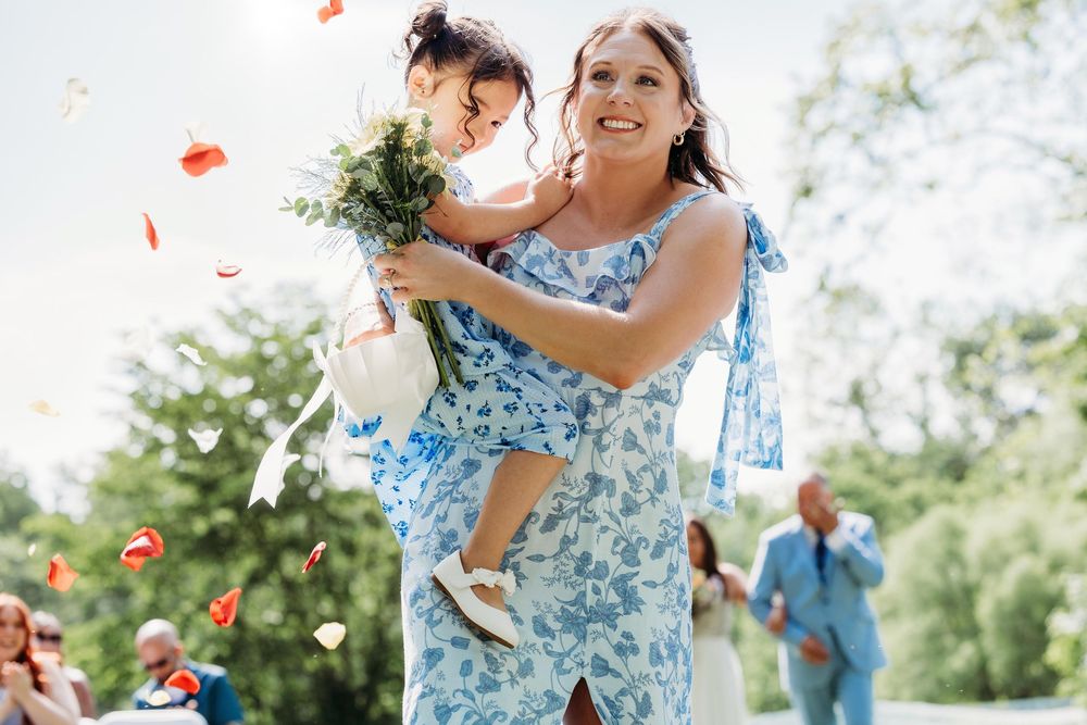 Woman in blue floral dress holds flower girl tossing rose petals at outdoor wedding ceremony.