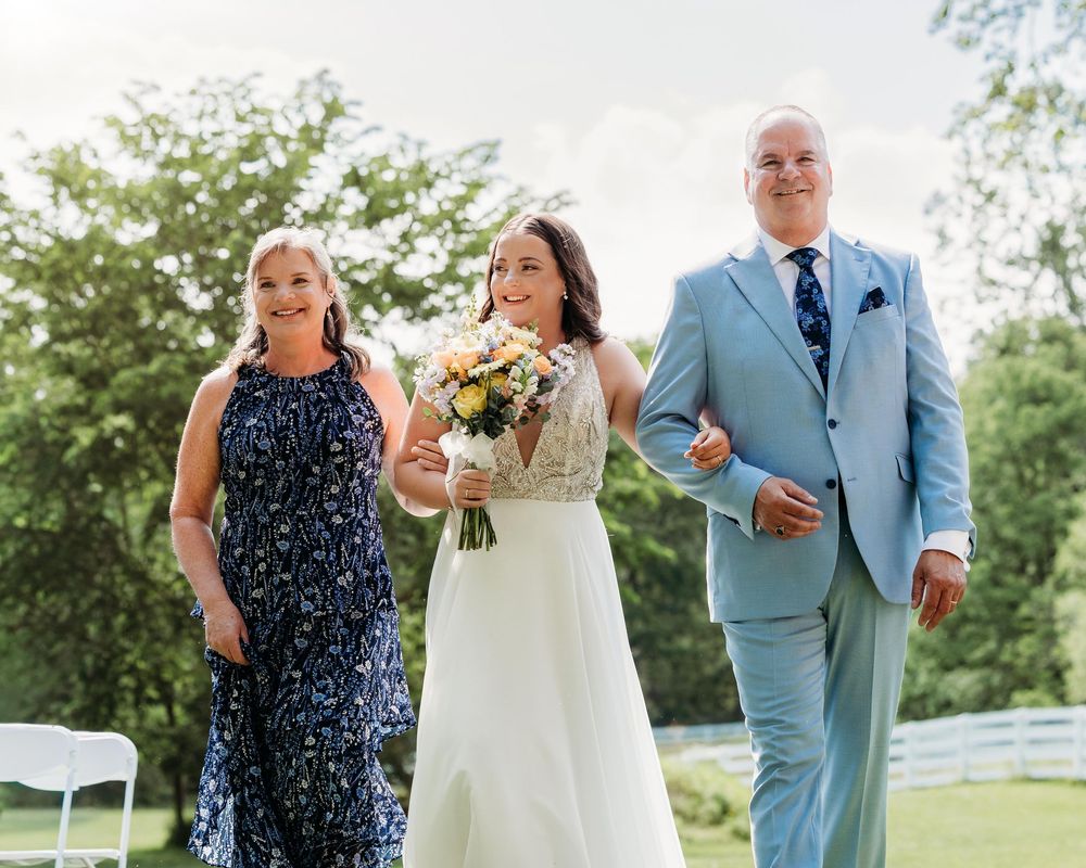 Bride in white gown holding bouquet walks down aisle flanked by parents at outdoor wedding ceremony.