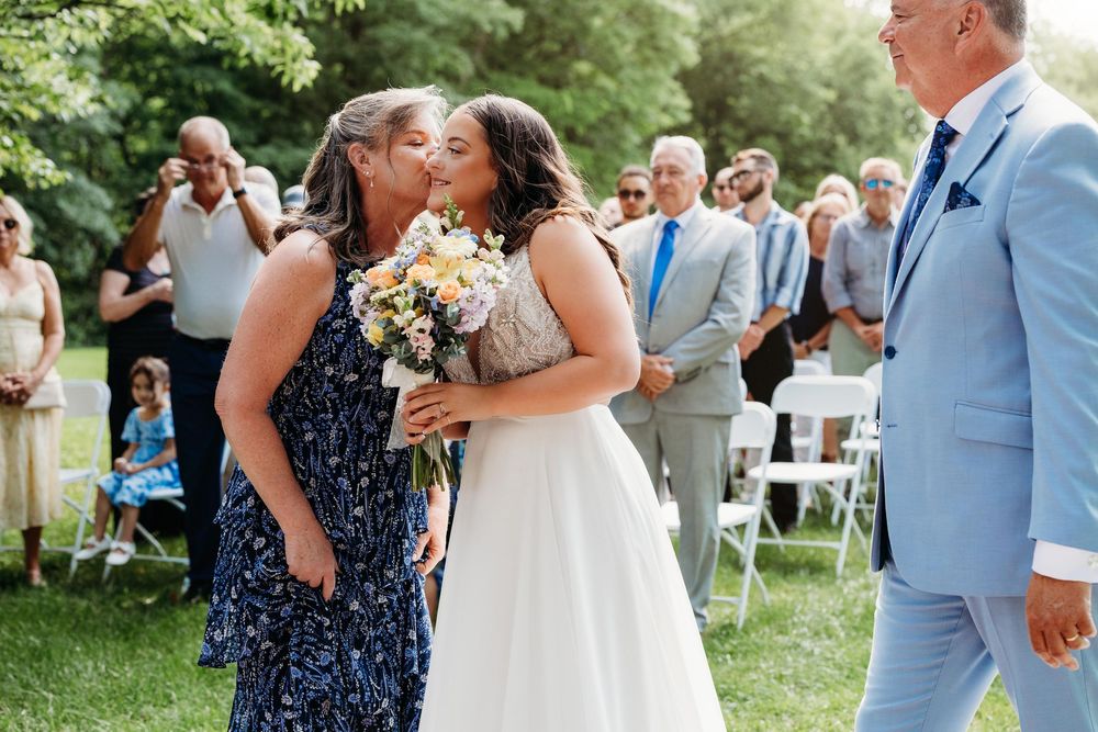 Bride receives a kiss from a guest while walking down the aisle at an outdoor wedding ceremony.