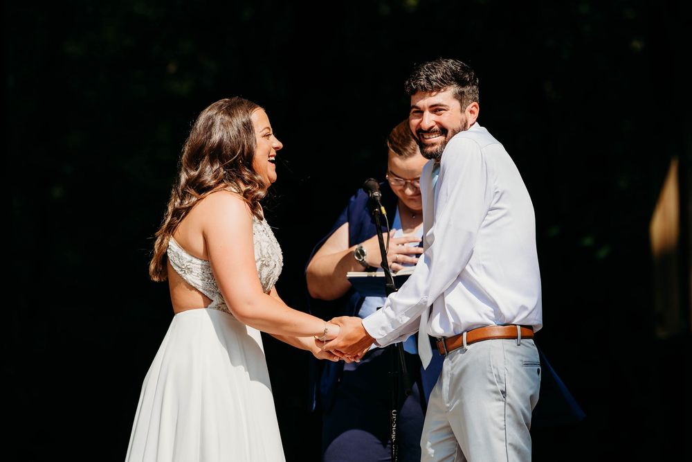 Joyful bride and groom holding hands and laughing during outdoor wedding ceremony with officiant in background.