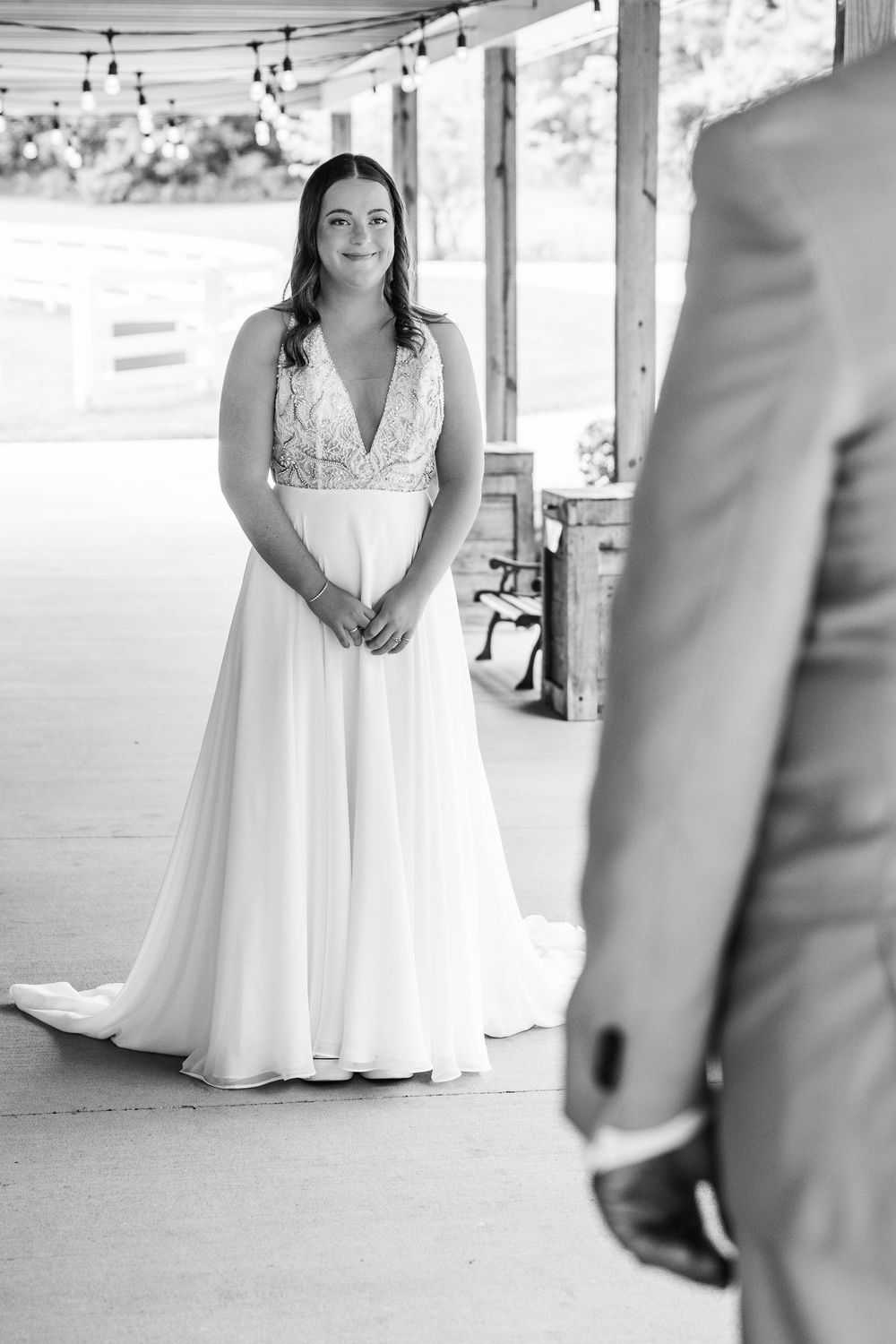 Black and white photo of a bride in a elegant gown smiling during a first look at an outdoor wedding venue.