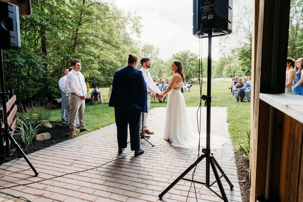 Outdoor wedding ceremony on a brick patio with bride and groom holding hands before an officiant, guests seated in background.