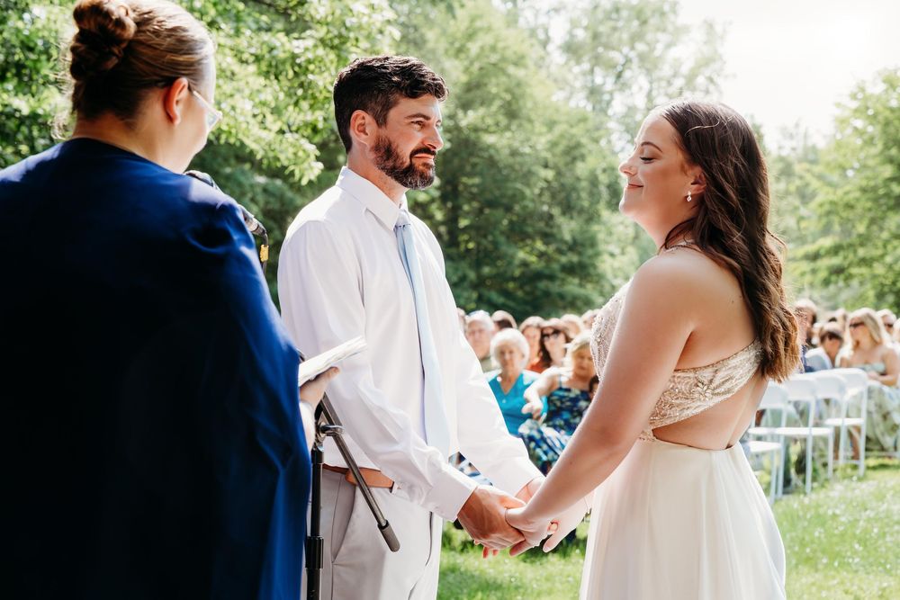 Bride and groom holding hands during outdoor wedding ceremony with officiant and seated guests in background.