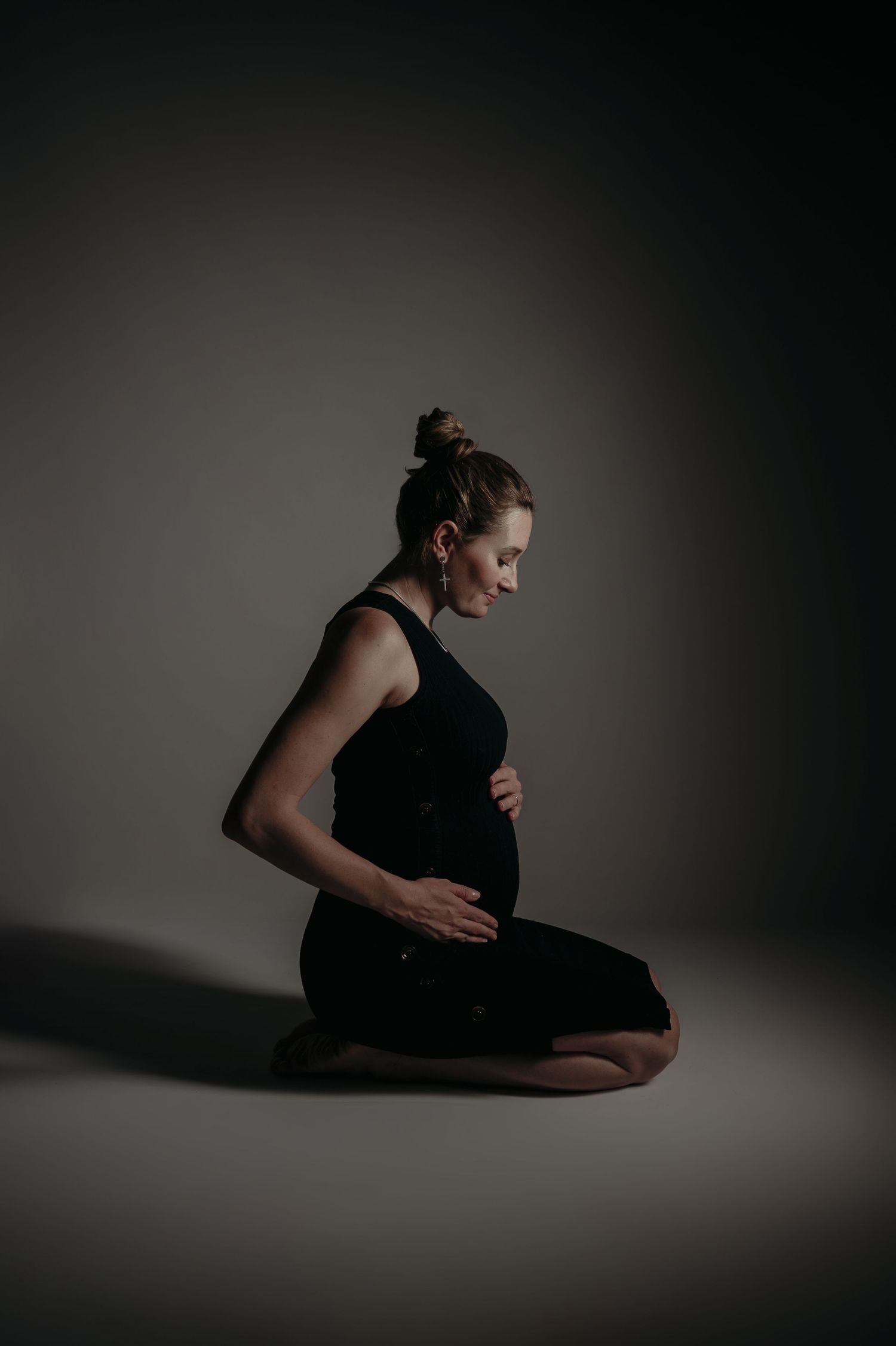 Pregnant woman in black dress sitting cross-legged on floor, cradling her baby bump in a moody studio setting.