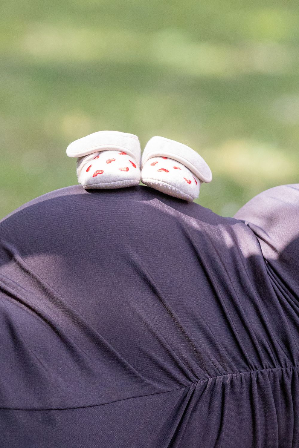 Paar witte babyschoentjes met rode hartjes staat op een zwangere buik in een donkere jurk.