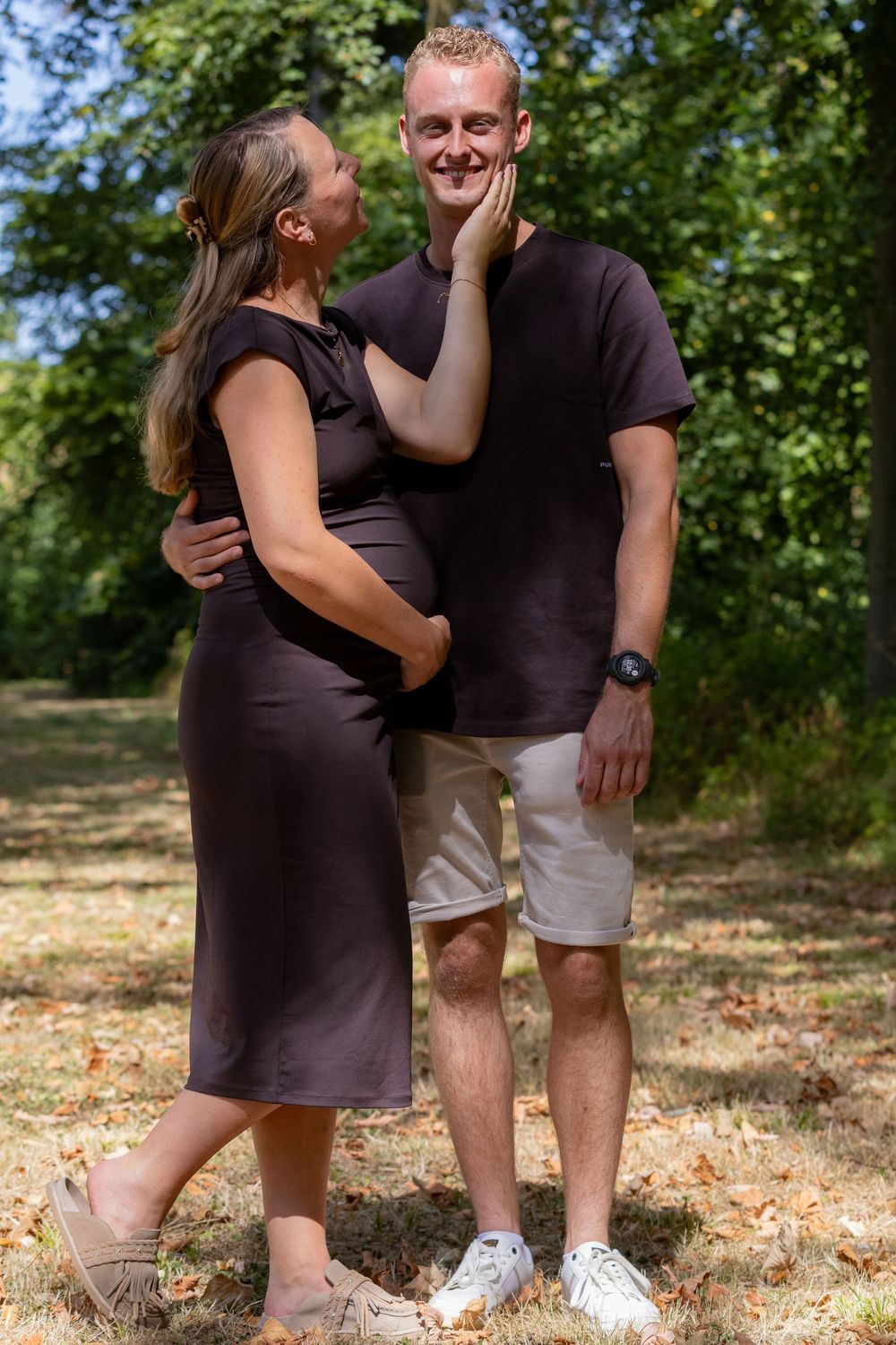 Zwangere vrouw in een donkere jurk kust haar glimlachende partner op de wang tijdens een fotoshoot in het bos.
