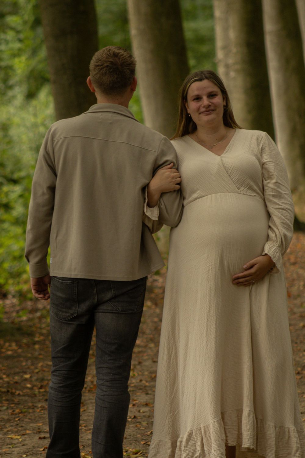 Glimlachende zwangere vrouw in een lange crèmekleurige jurk loopt hand in hand met haar partner door het bos.