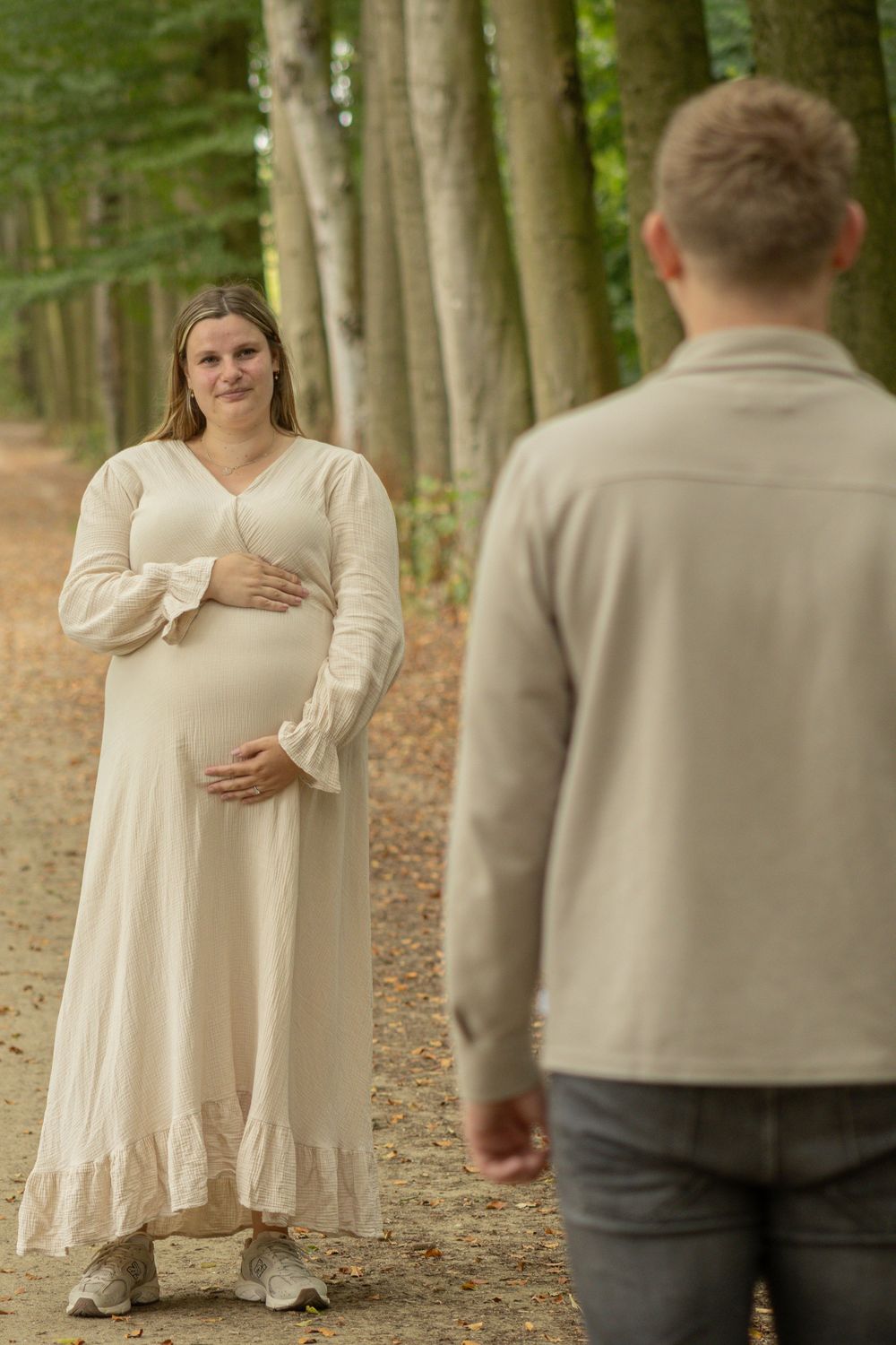 Zwangere vrouw in een lange crèmekleurige jurk kijkt glimlachend naar haar partner op een bospad.