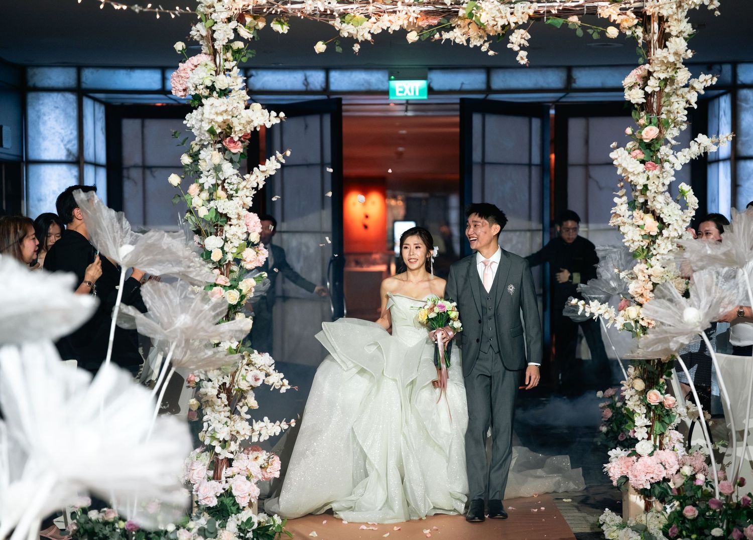 A couple in wedding attire share a playful moment together on a lawn with white buildings in the background.