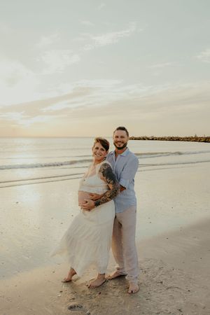 Pregnant couple embracing on beach at sunset, woman in white dress, man in blue shirt with tattoos.