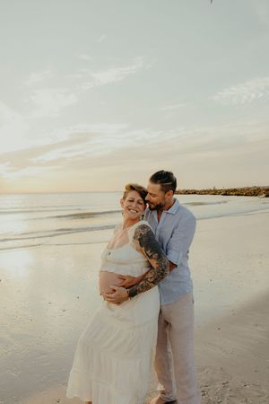 Pregnant couple embracing on beach at sunset, woman in white dress with tattoos, man in blue shirt holding her baby bump.