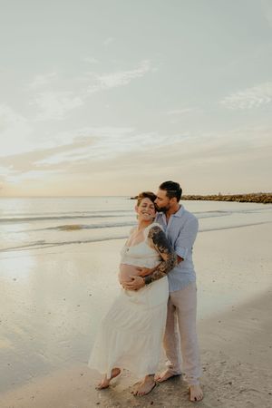 Couple embracing on beach at sunset, pregnant woman in white dress with tattooed partner holding her belly.