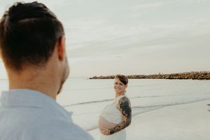 Pregnant woman with tattoos smiles on beach while partner stands in foreground during maternity photo shoot.