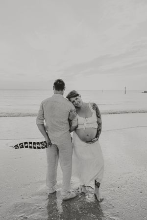 Black and white beach maternity photo of pregnant woman in white dress leaning on partner, waves in background.