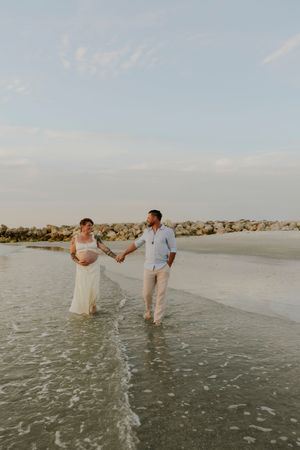 Expecting couple holds hands walking along shallow beach waves at sunset in white and blue outfits.