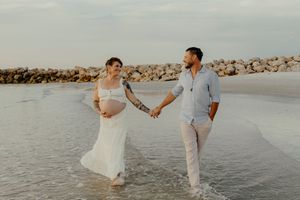 Pregnant couple holding hands walking in ocean waves during a beach maternity photo shoot at sunset.