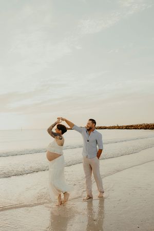 Pregnant couple dancing on beach at sunset, woman in white dress with baby bump, man in light blue shirt and white pants.
