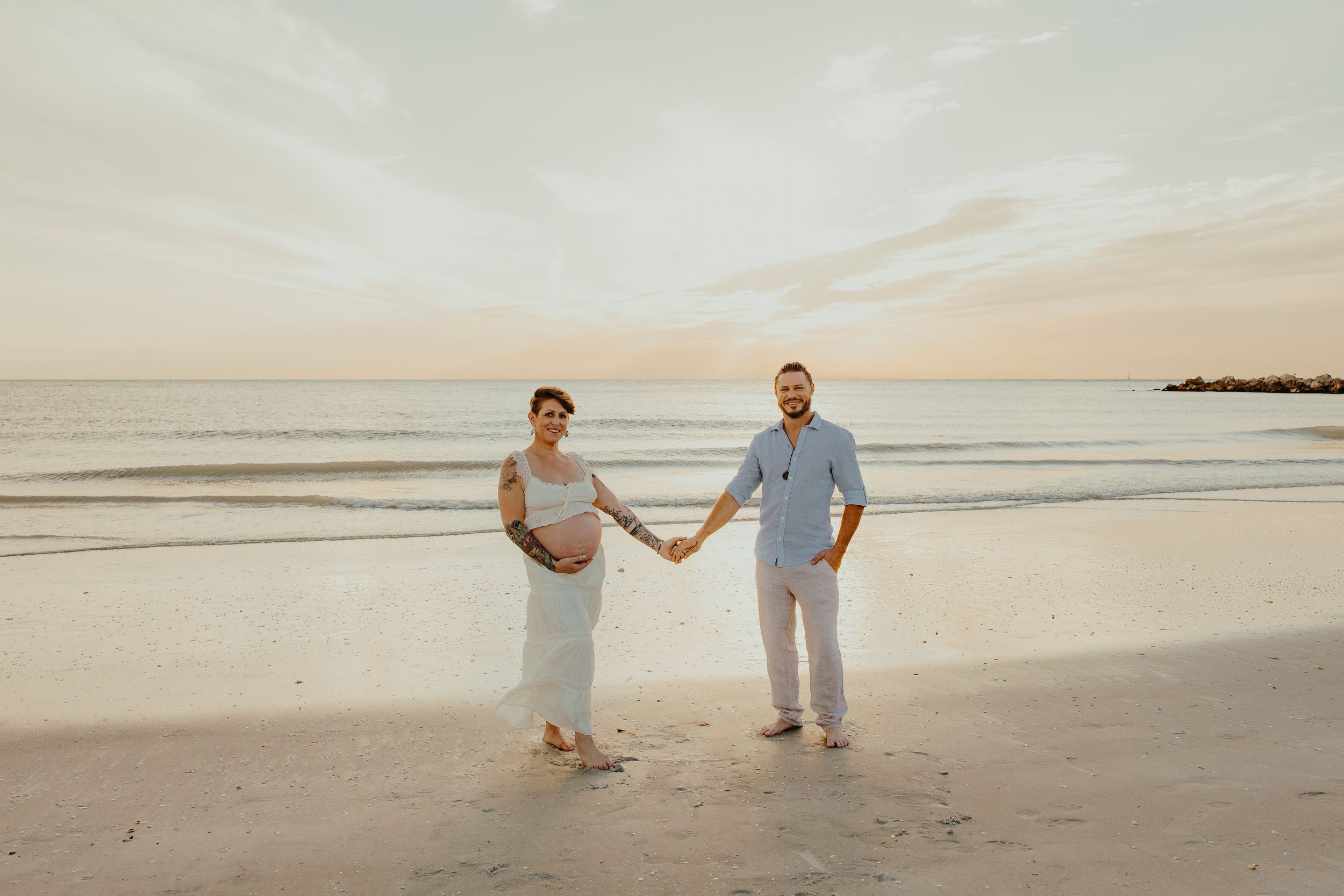 Pregnant couple holding hands on a sandy beach at sunset, woman in white dress showing baby bump.