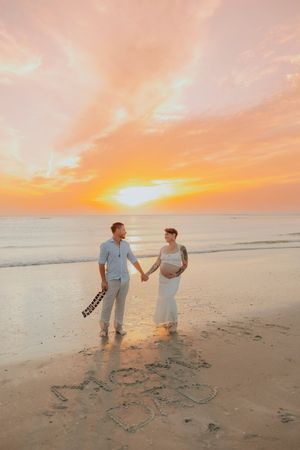 Pregnant couple holds hands on beach at sunset with MOM and DAD written in sand, golden sky reflecting on ocean waves.