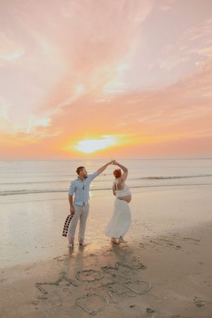 Pregnant couple dancing on beach at sunset, man holding guitar, words written in sand, romantic golden sky backdrop.