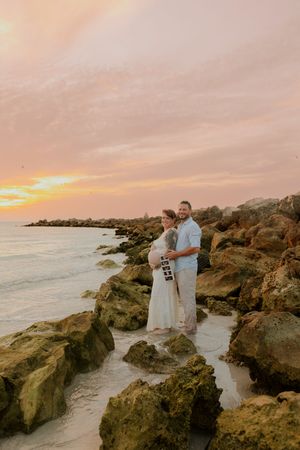 Couple holding ultrasound photos on rocky beach at sunset, maternity photography session.