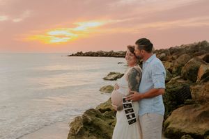 Couple embracing at sunset on rocky beach, woman holding ultrasound photos, announcing pregnancy.