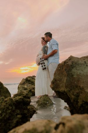Pregnant couple embracing on rocky shoreline at golden sunset, holding ultrasound images.