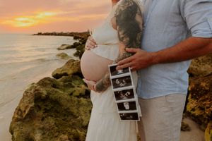 Pregnant couple embracing at sunset beach, holding ultrasound photos by rocky shore.