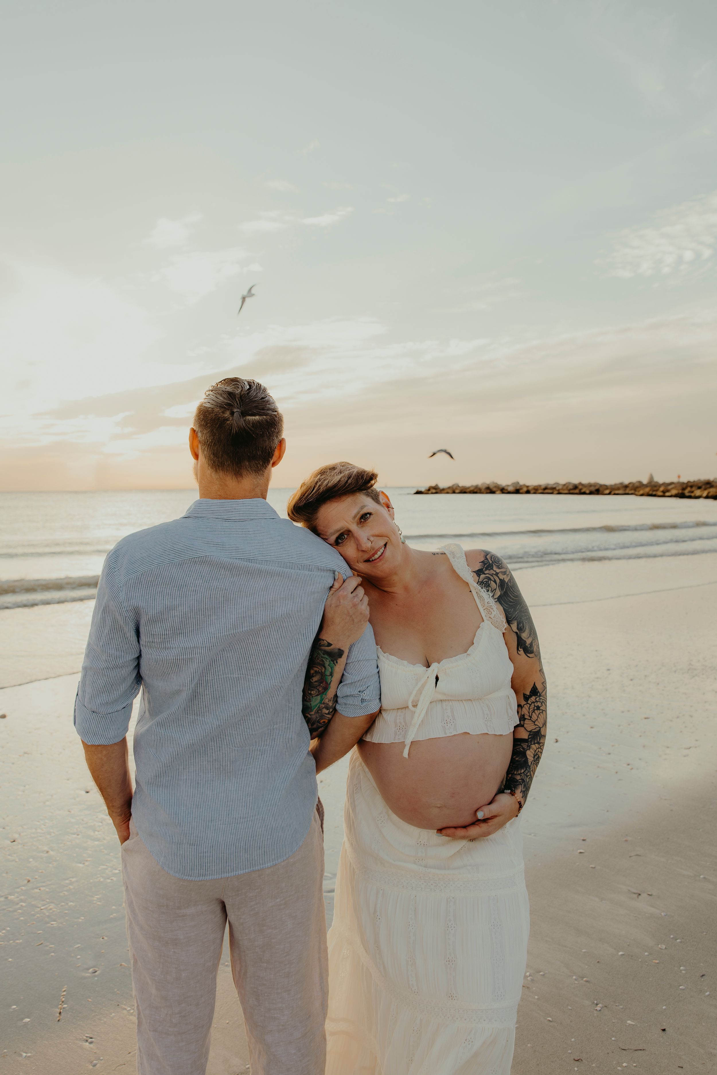 Pregnant woman with tattoos smiles at camera on beach during sunset while partner stands beside her.