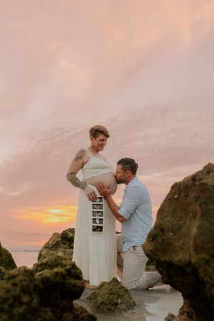 Man kneels kissing pregnant woman's belly holding ultrasound photos on rocky beach at colorful sunset.