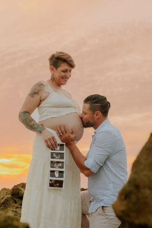 Pregnant woman in white dress holds ultrasound photos as partner kisses her baby bump during golden sunset beach maternity shoot.