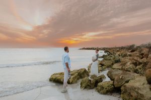 Expecting couple stands on rocky shoreline during vibrant pink and orange sunset in a romantic beach maternity photo session.