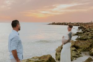Pregnant couple sharing a romantic moment on rocky shoreline during a golden sunset over the ocean.
