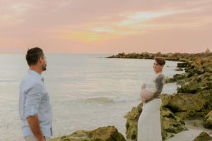 Couple standing on rocky shore at sunset, woman in white dress cradling baby bump, man in light blue shirt facing her.