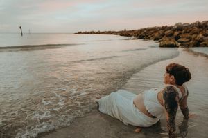 Tattooed pregnant woman in white dress lying on sandy beach shore at sunset with rocky jetty in background.
