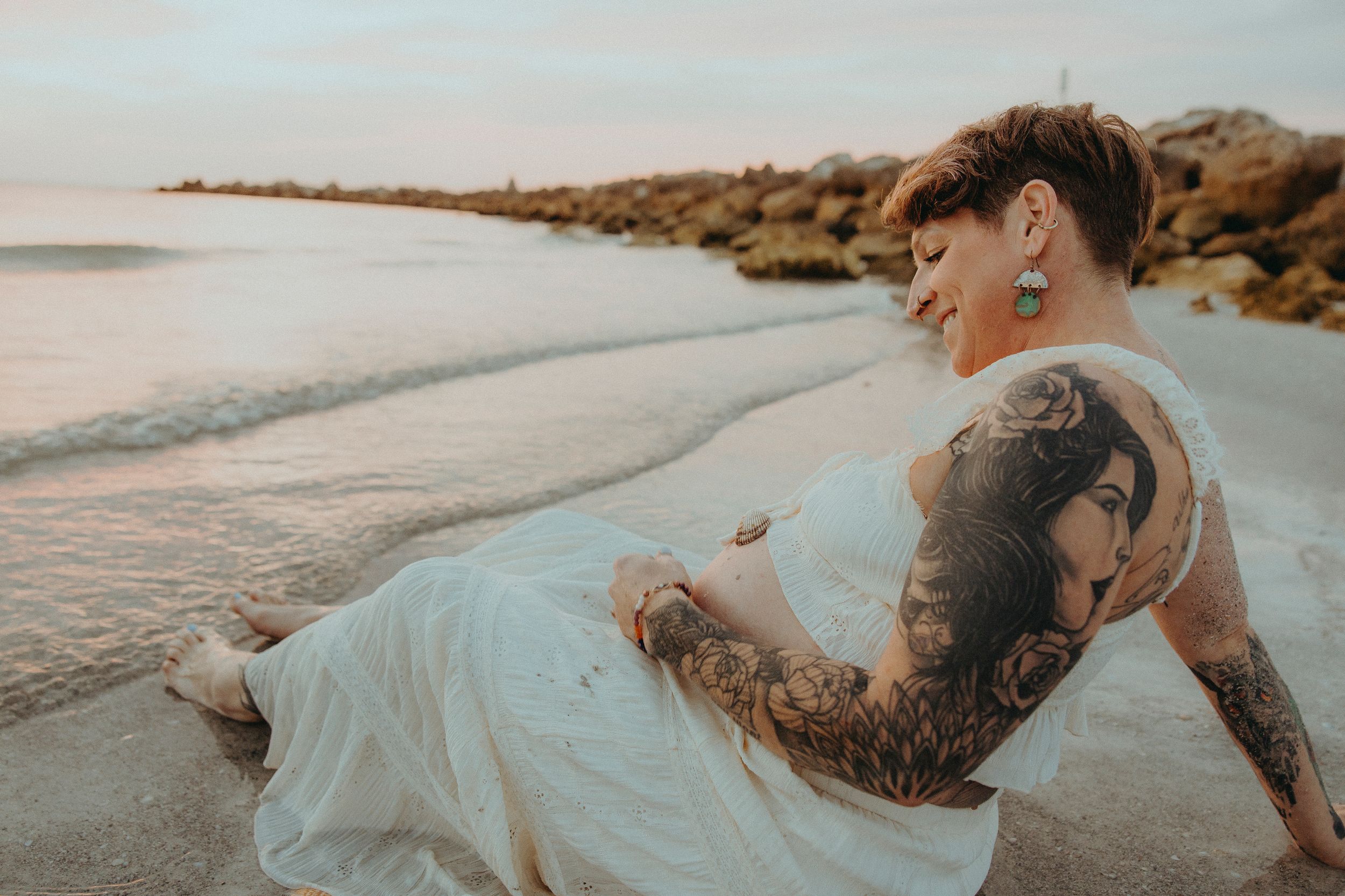 Tattooed bride in white dress sitting on sandy beach at sunset with rocky shoreline in background.