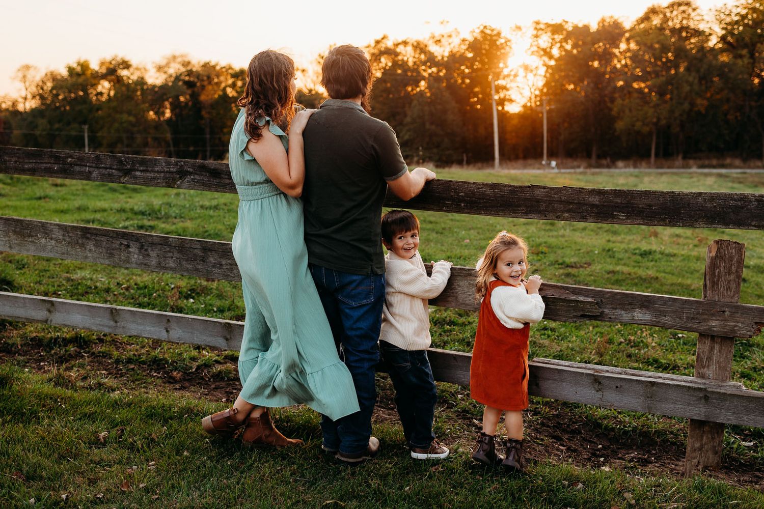 A family of four in the countryside as the sunsets. the couple gazes in the distance, and two children are looking back and smiling.