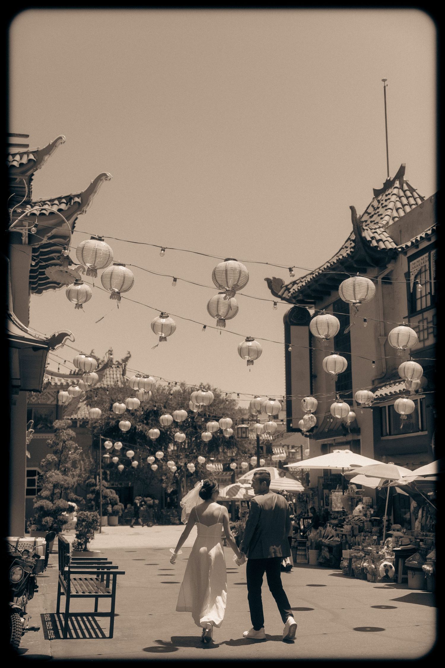 Couple walks hand-in-hand through Chinatown plaza with hanging lanterns and traditional architecture in sepia tone.