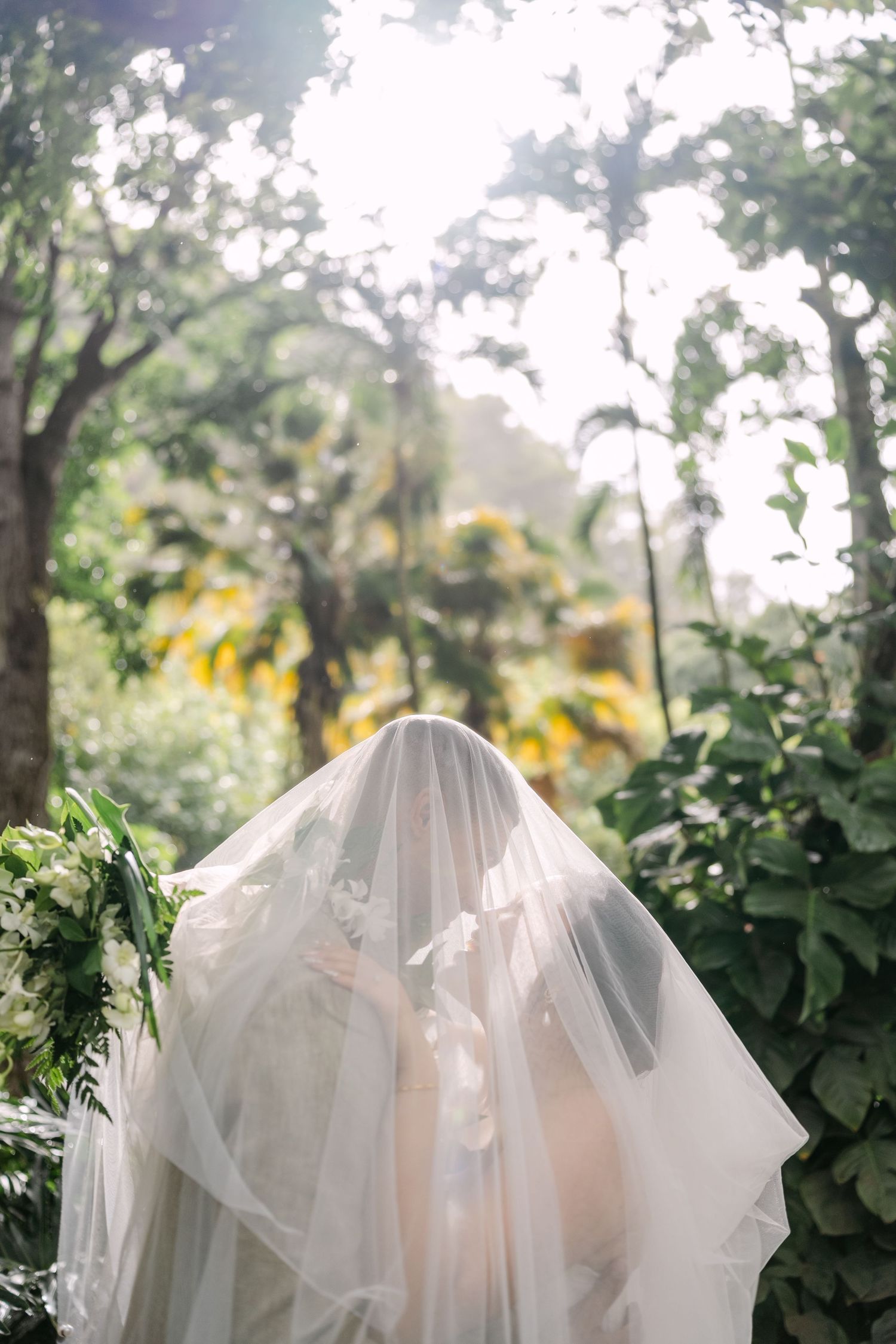 Bride in white wedding gown and flowing veil stands in lush tropical garden with sunlight filtering through trees.