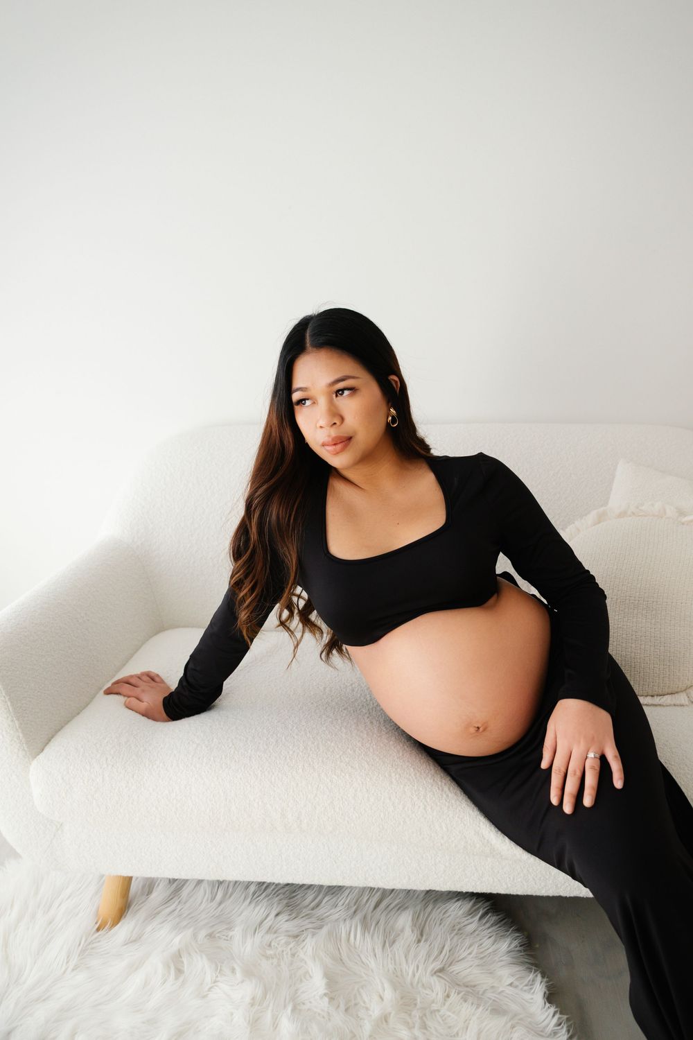 Pregnant woman in black crop top and skirt sits on white sofa, showing bare bump in bright studio.