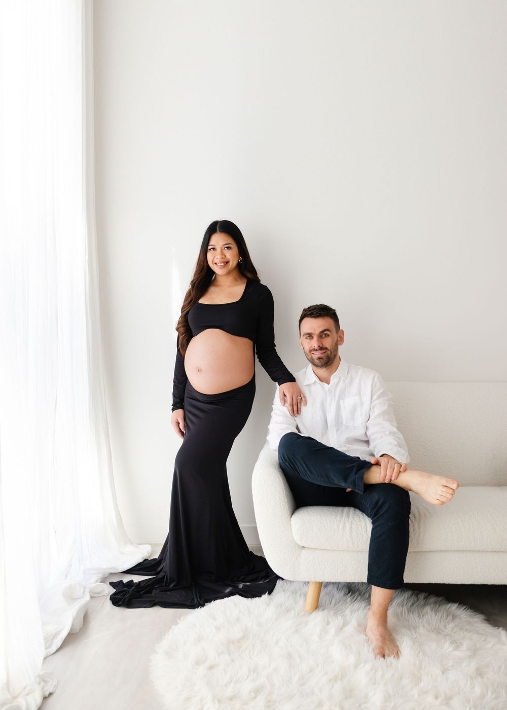 Pregnant woman in black gown poses with man in white shirt beside cream sofa in bright white studio maternity photo.